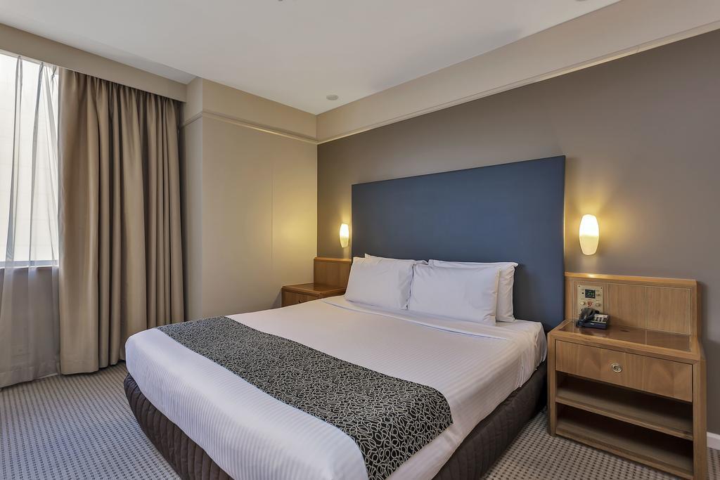 Stamford Plaza Adelaide Hotell Exteriör bild The photo shows a modern hotel room. It features a double bed with a white duvet and a decorative grey and black throw. The headboard is a solid dark color. There are two bedside tables on either side of the bed, each with a lamp. Natural light enter